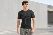 Männliches Model trägt ein schwarzes, körpernahes Funktionsshirt mit strukturierten Ärmeln und grafischen Details auf der Vorderseite. Es steht entspannt vor einer modernen hellen Betonarchitektur im Außenbereich und trägt dazu hellolivfarbene Sportshorts.